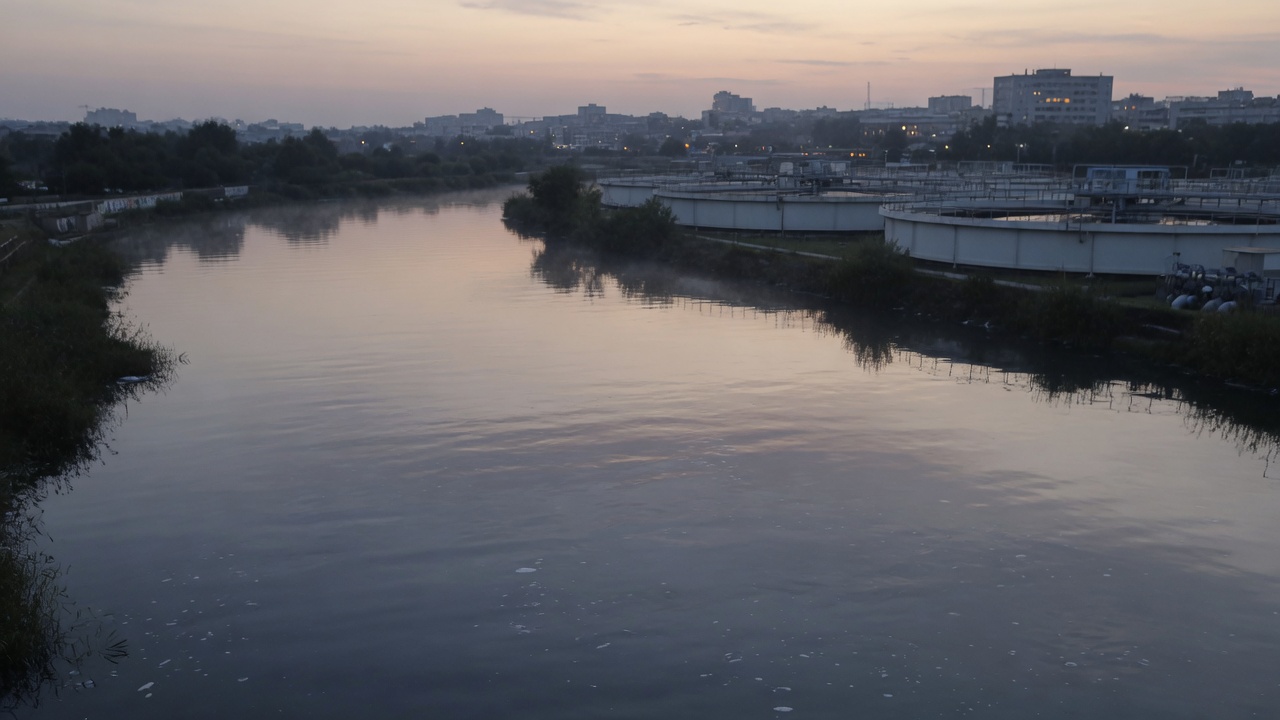 A wide river at dusk with distant industrial water-treatment forms on the horizon